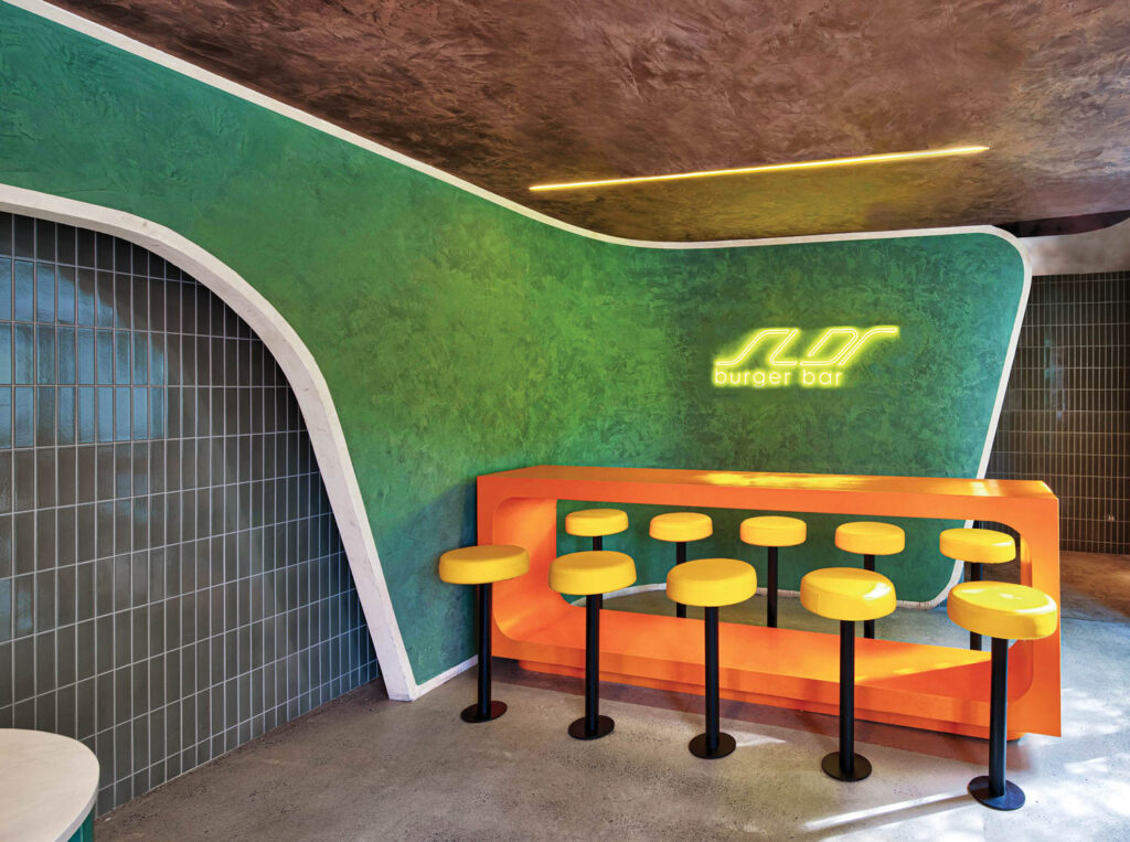 Neon yellow signage pops against a green curved wall with an orange dining counter and bright yellow stools.