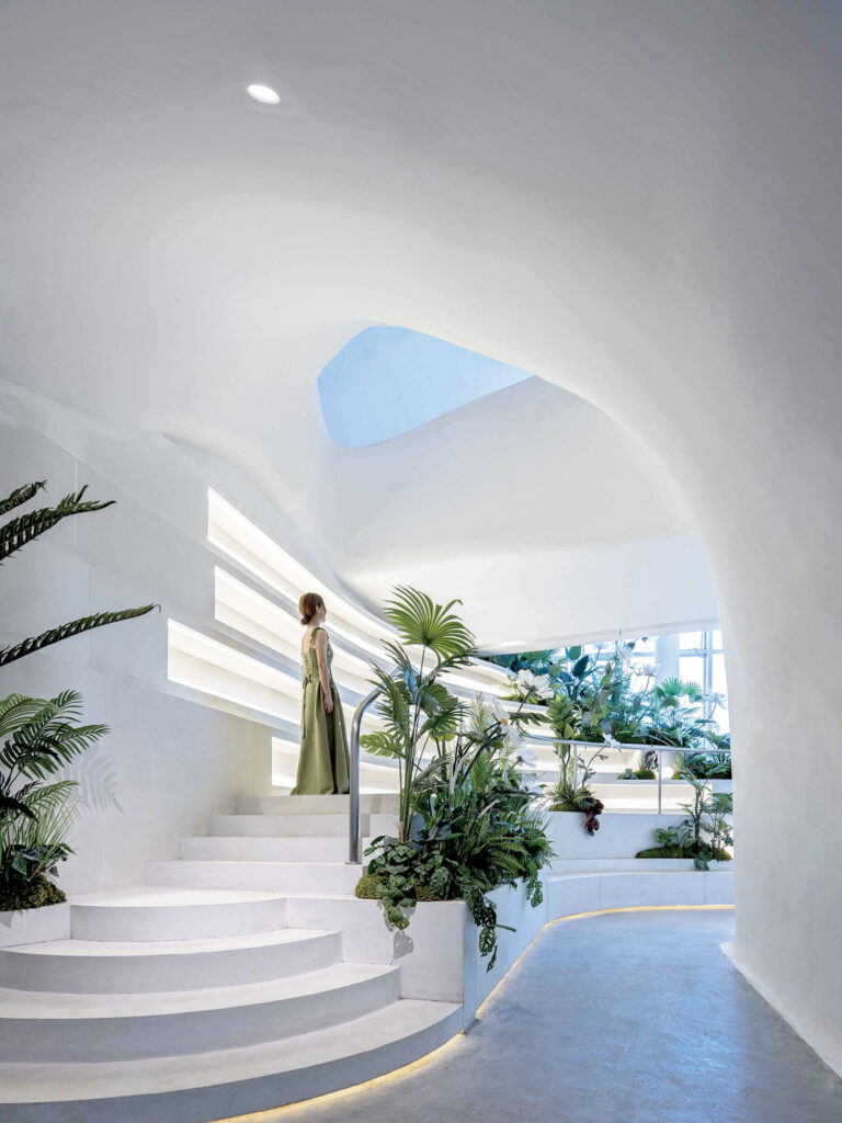 a woman in green walks up stairs overlooking the dining room of white walls and greenery