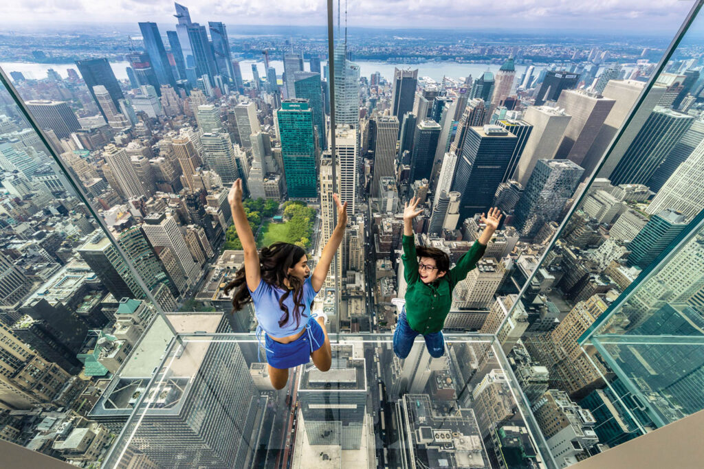 2 people jump in the air inside Summit One Vanderbilt, with NYC visible in the background