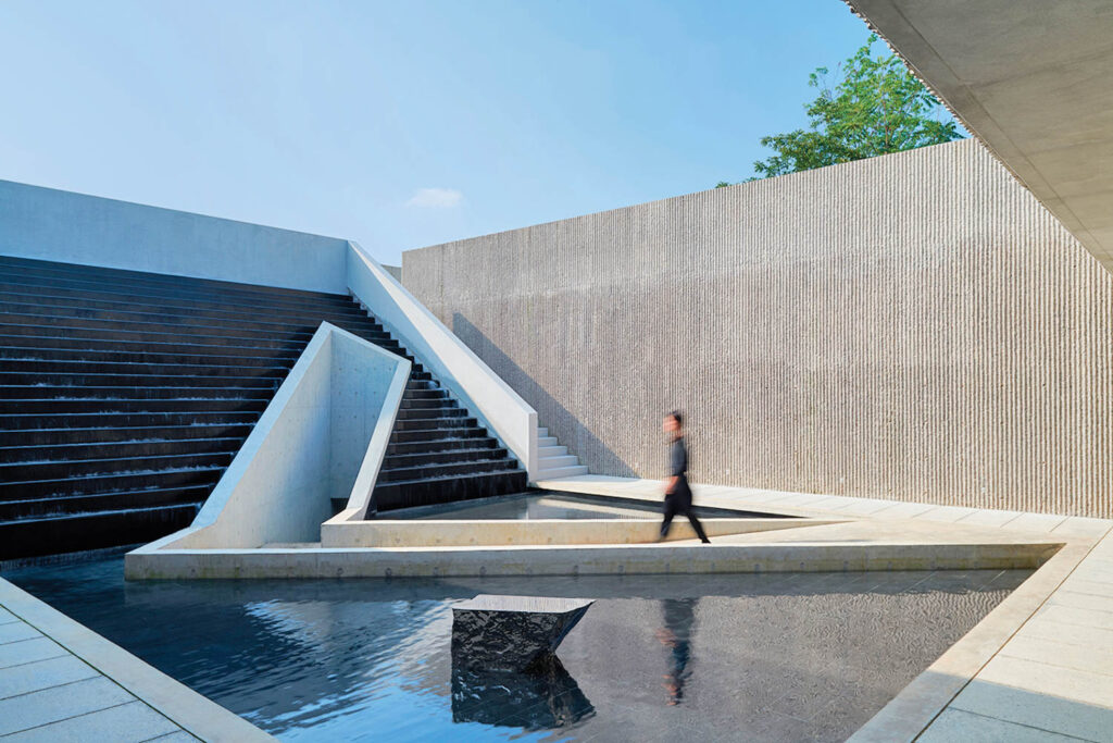 a man walks toward stairs on an architectural building with a pond in front