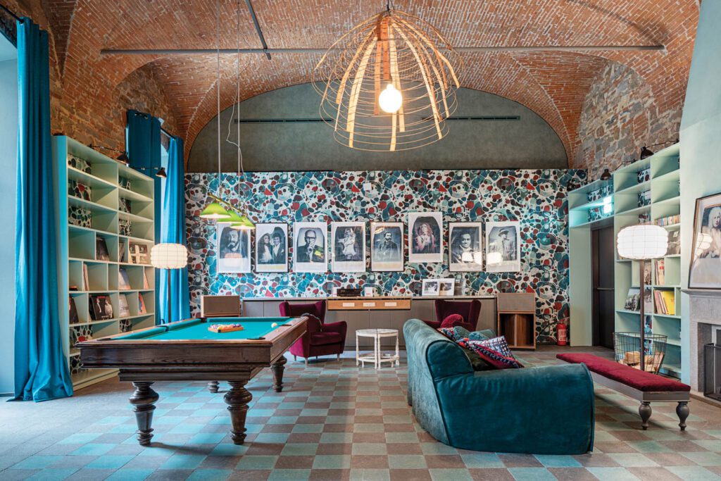 Composite-stone tile flooring, custom vinyl wallcovering, and a hoop-skirt frame used as a pendant fixture join a billiard table and a display of Italian celebrity portraits in the Sala delle Celesti Armonie, or music room.